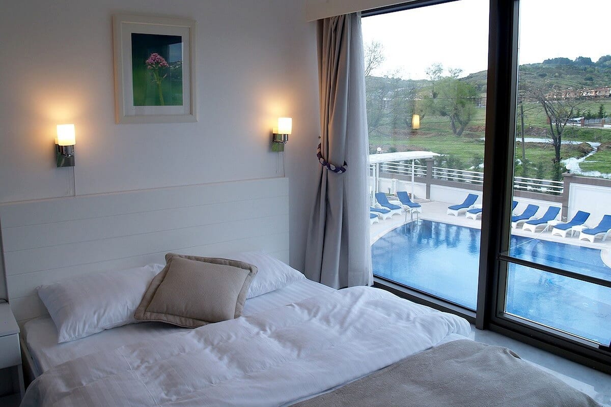 A hotel room with a bed, two wall-mounted lights, a framed picture, and a large window overlooking a pool and outdoor area.