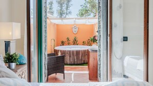A room with a bed, a small table with a lamp, and a view of an outdoor area with a wooden fence and plants.