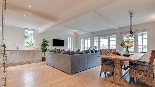 A spacious living area with a grey sofa, a dining table with wicker chairs, and a kitchen with white cabinets.