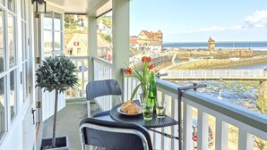 A balcony with a table, chairs, a potted plant, and a view of the sea and buildings.