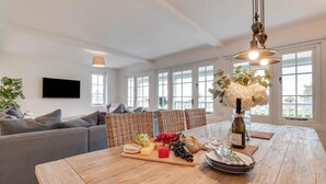 A modern living room with a grey sofa, wicker chairs, a wooden dining table with a fruit platter, and a wine bottle.