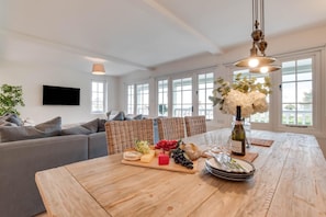 Un salon moderne avec un canapé gris, des fauteuils en osier, une table à manger en bois sur laquelle est disposée une corbeille de fruits, et une bouteille de vin.