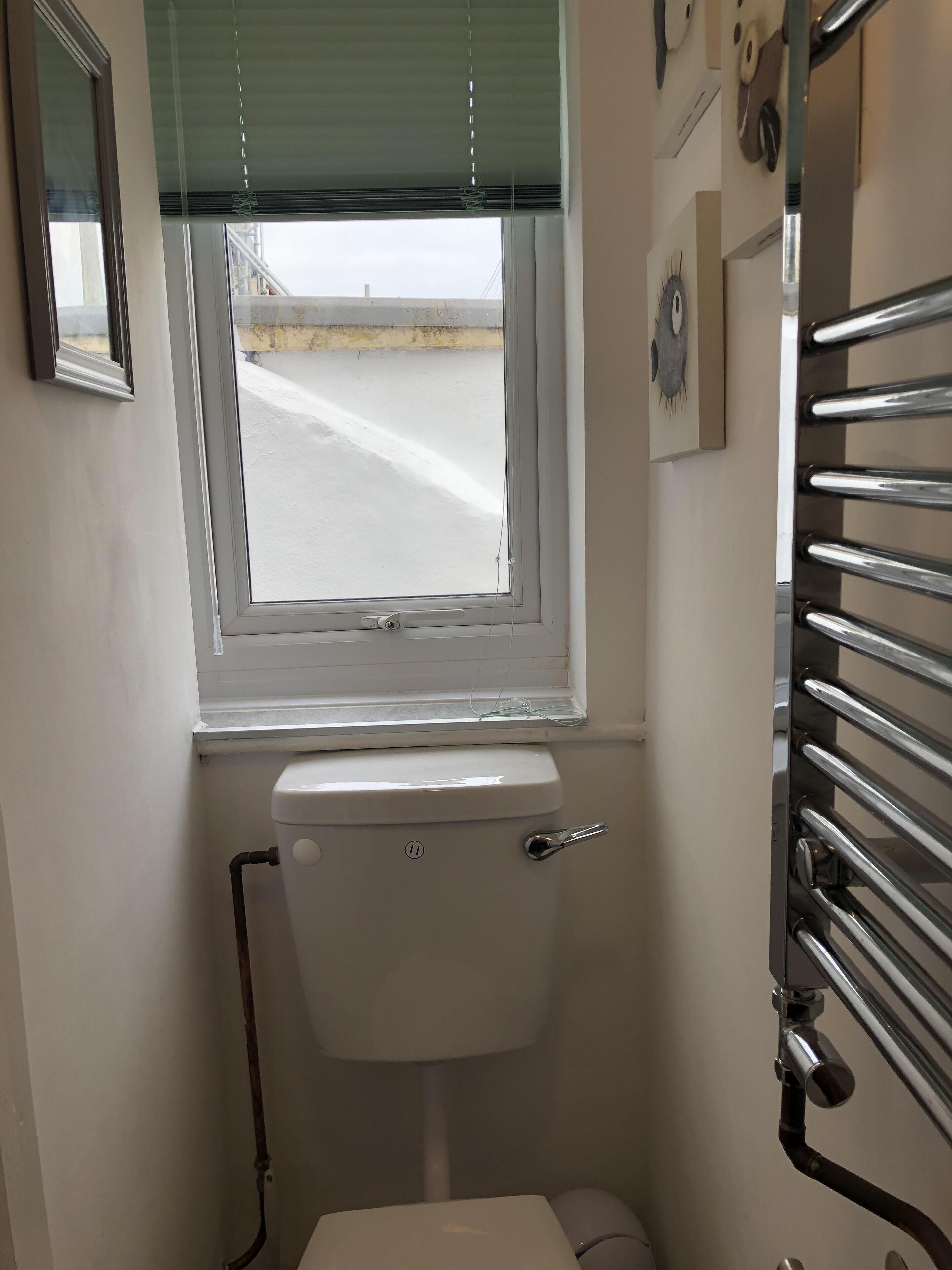 Une petite salle de bain comprenant des toilettes, une fenêtre avec des stores, un porte-serviettes et une horloge fixée au mur.