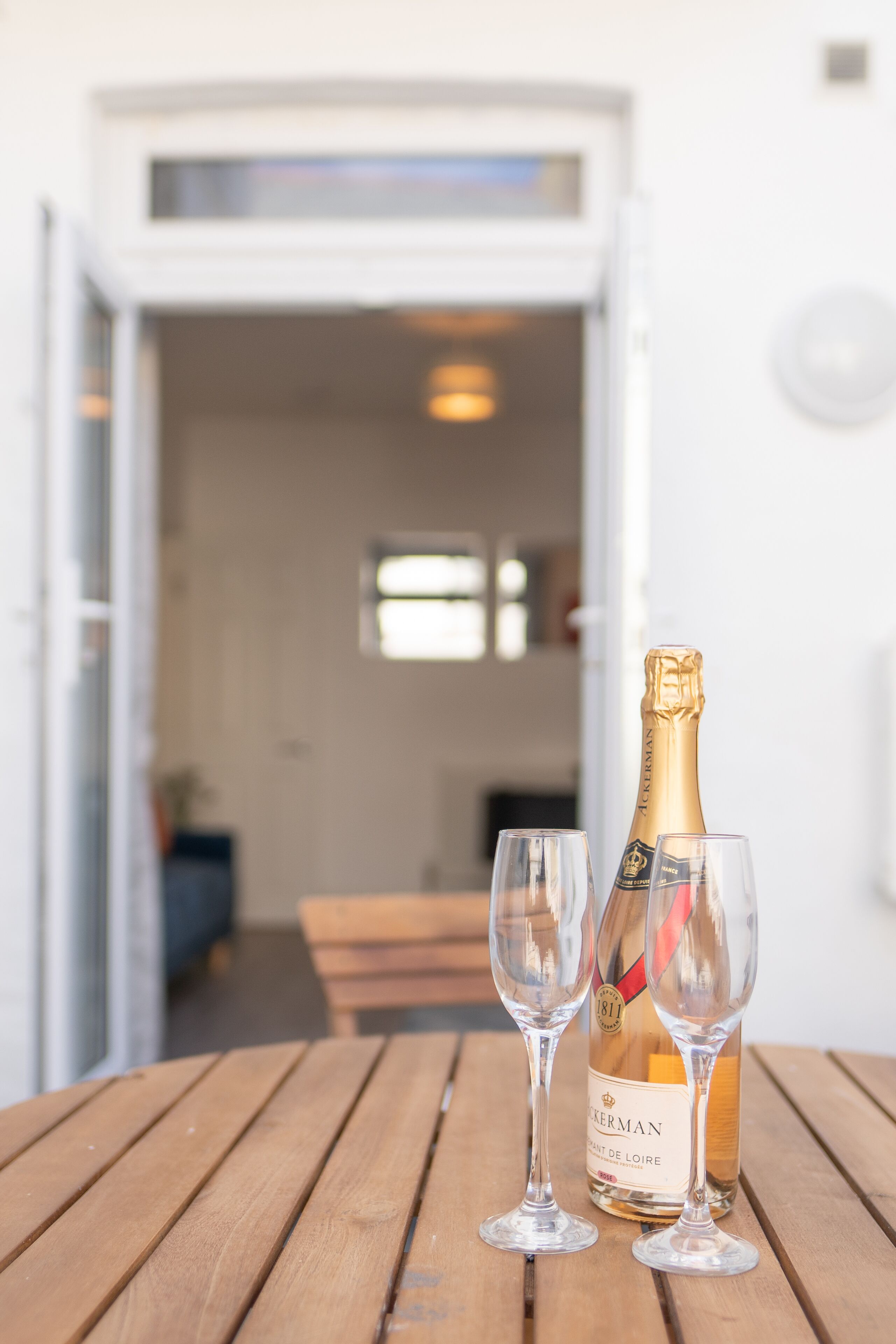 A bottle of champagne and two glasses on a wooden table, with a doorway leading to a room in the background.