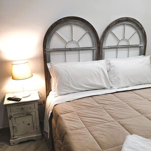 A bed with a beige quilt, two white pillows, and a nightstand with a lamp and a remote control. - Art Flat (Naples)