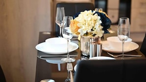 A dining table set with white plates, silverware, and wine glasses, accompanied by a vase of white and dark blue flowers. - Livestay - Modern Spacious 2 Bed 2 Bath Apartment (Slough)