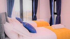A hotel room with two beds, each with a different colored bedspread, a grey headboard, and a window with blue curtains. - Livestay - Modern Spacious 2 Bed 2 Bath Apartment (Slough)