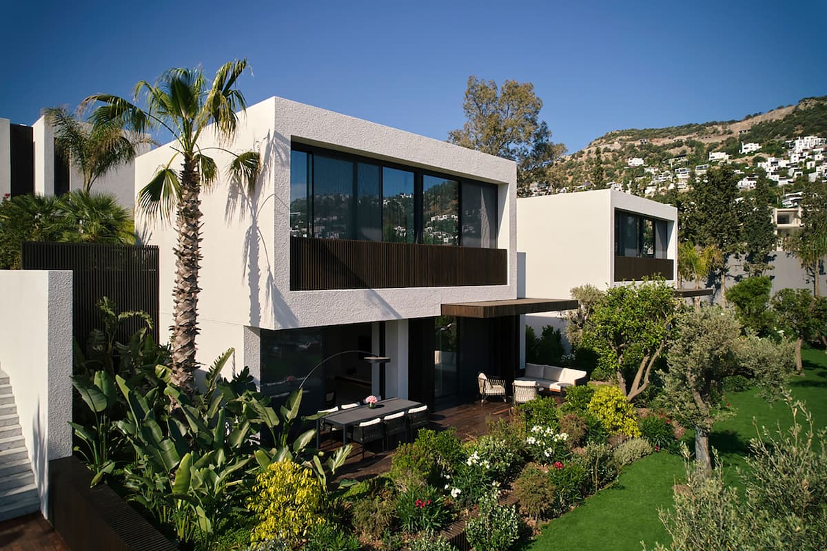 Une maison moderne à deux étages, dotée d’un patio, de grandes fenêtres et d’un jardin où poussent différentes espèces de plantes.