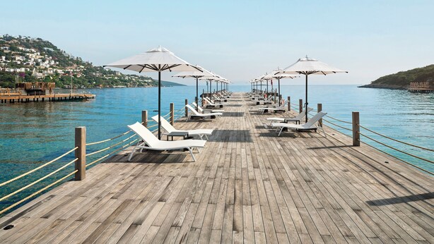 Un muelle de madera con sillones blancos y sombrillas, que se extiende hacia un mar tranquilo con una colina al fondo.