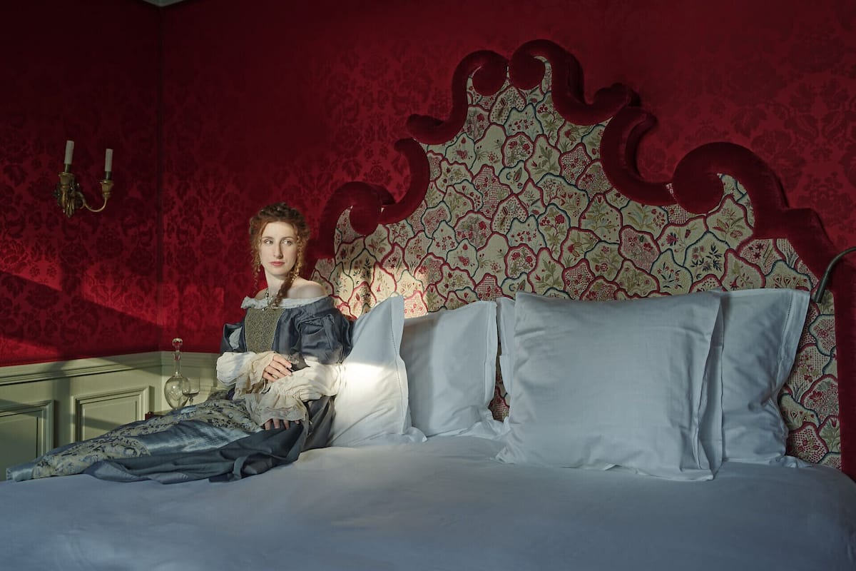 Un dormitorio con papel tapiz rojo con estampado, una cama blanca con muchas almohadas y un cabecero decorativo.