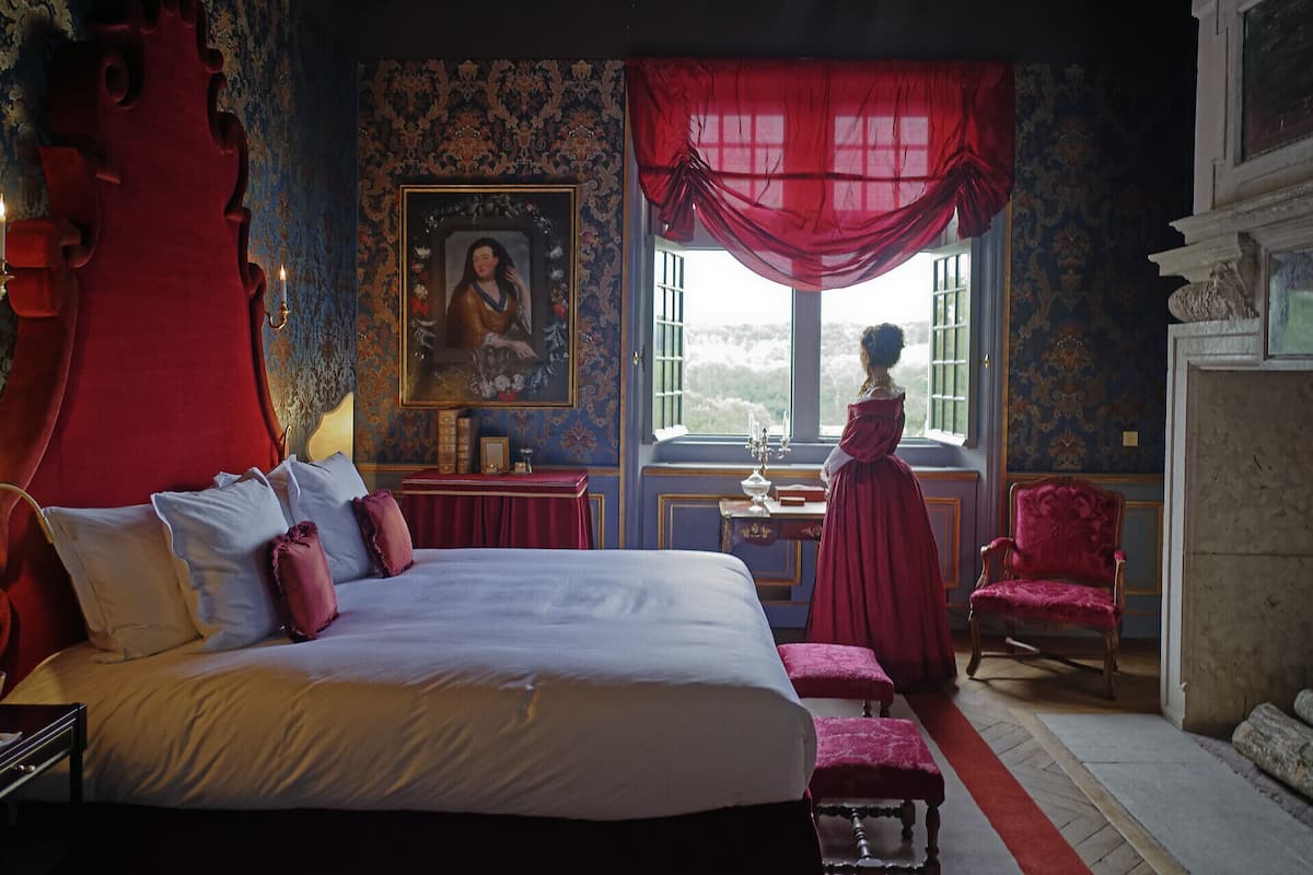Un dormitorio con una cama grande, un escritorio con silla, una ventana con cortinas rojas y una chimenea.