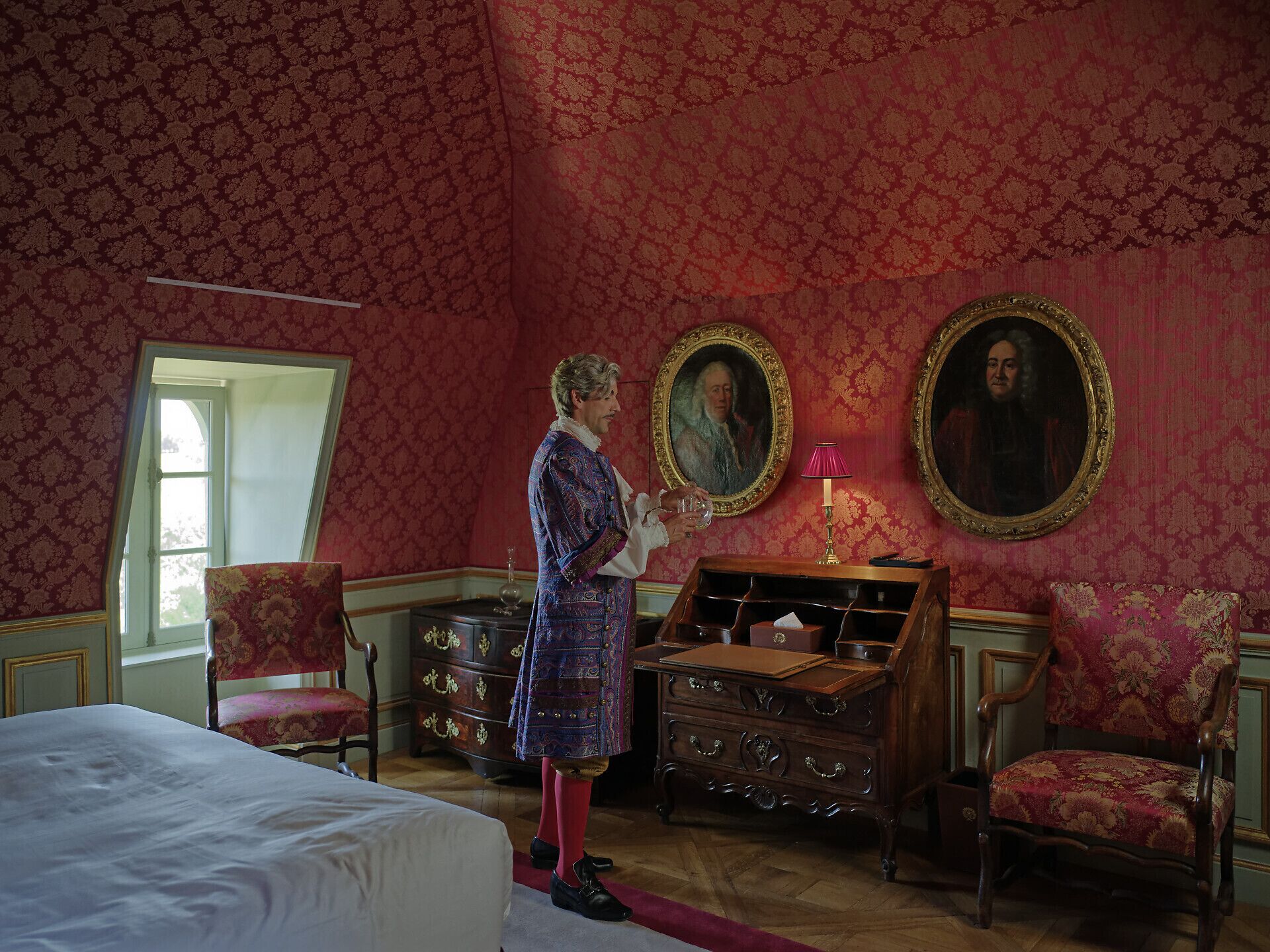 A room with a bed, a desk with drawers, two chairs, and two framed portraits on the wall.
