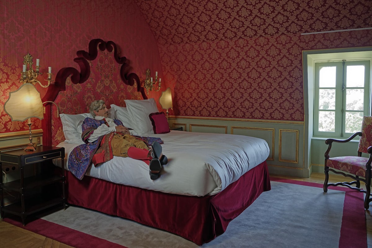 Un dormitorio con una cama grande, cabecera decorada, dos mesitas de noche con lámparas, una silla y un ventanal.