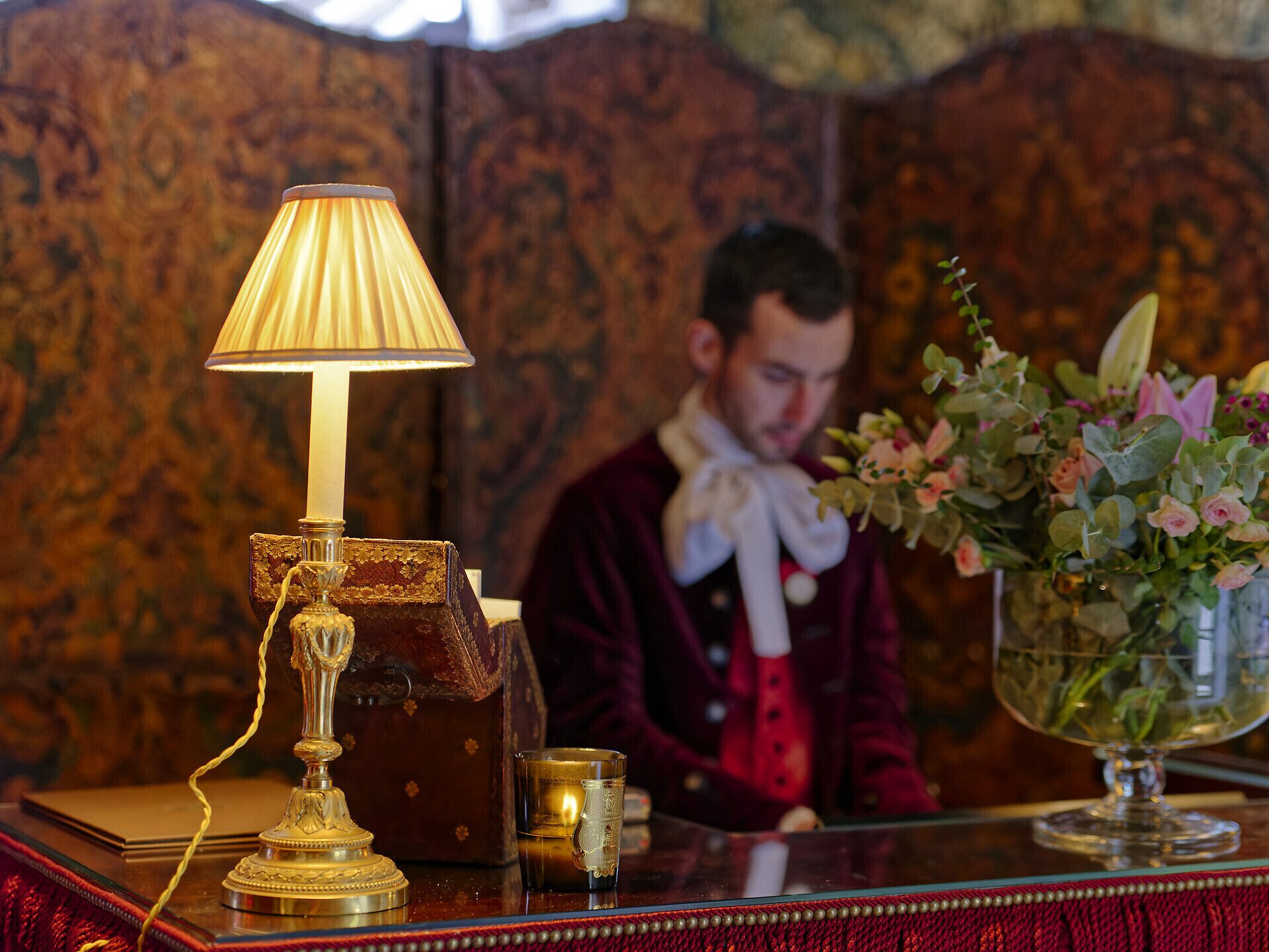 A table with a lamp, a candle, a glass, and a vase with flowers.