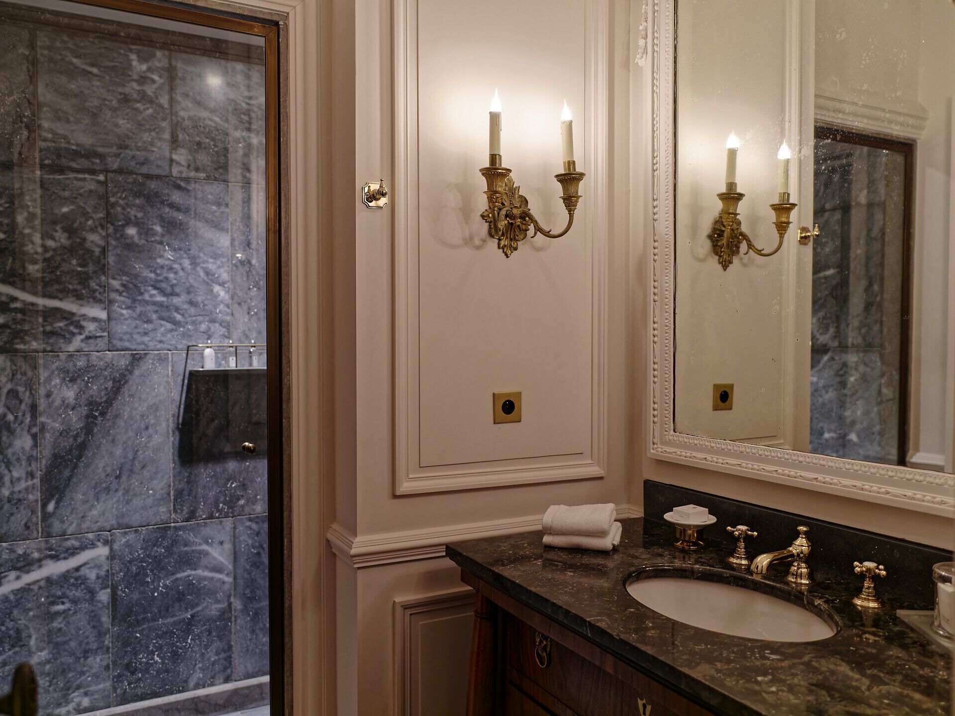 A bathroom with a marble wall, a sink with gold faucets, and a wall-mounted mirror.