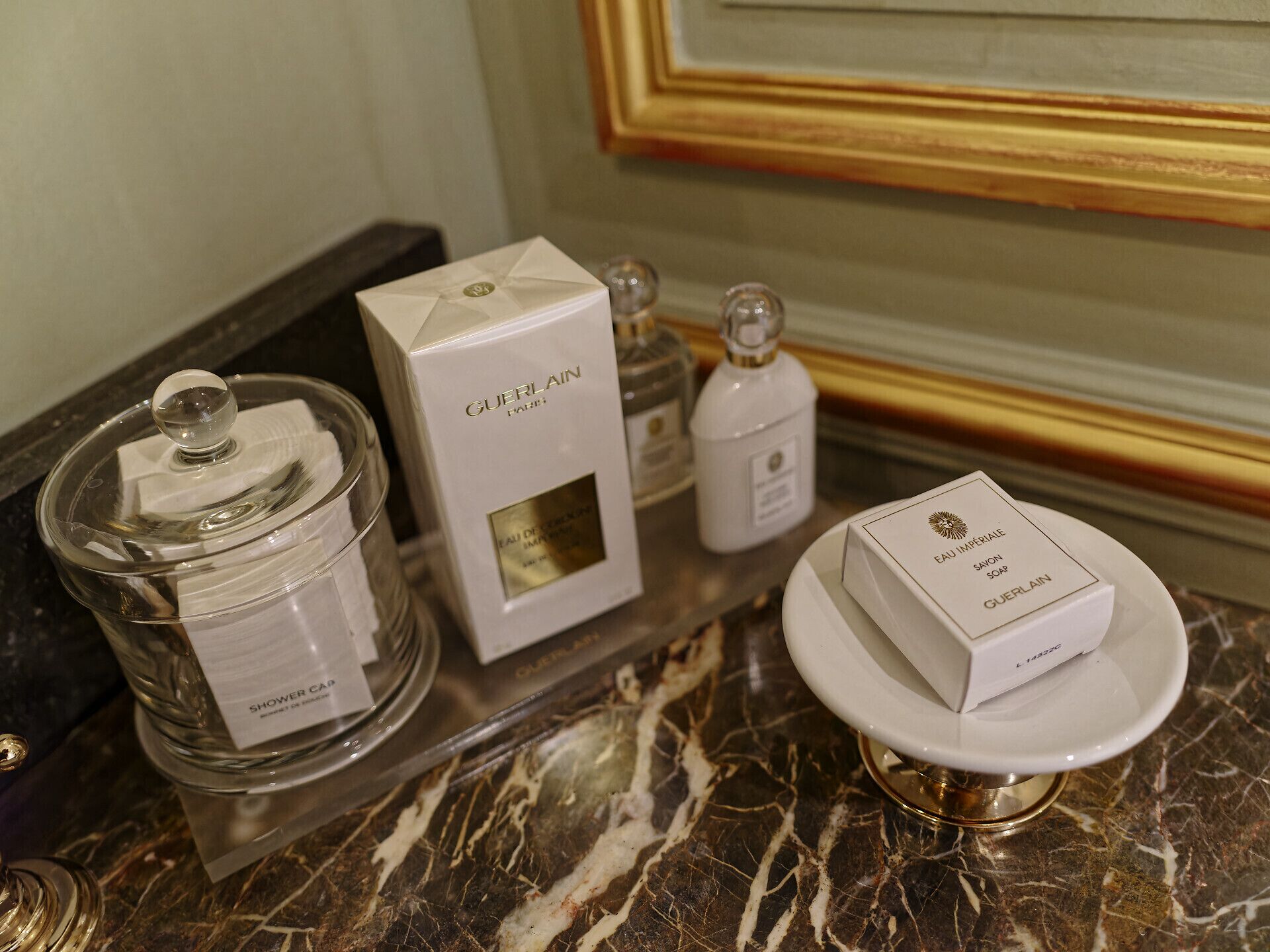 Un tocador de baño con encimera de mármol, un tarro de vidrio con tapa, una caja blanca con la etiqueta 'GUERLAIN PARIS' y una pequeña botella de jabón.