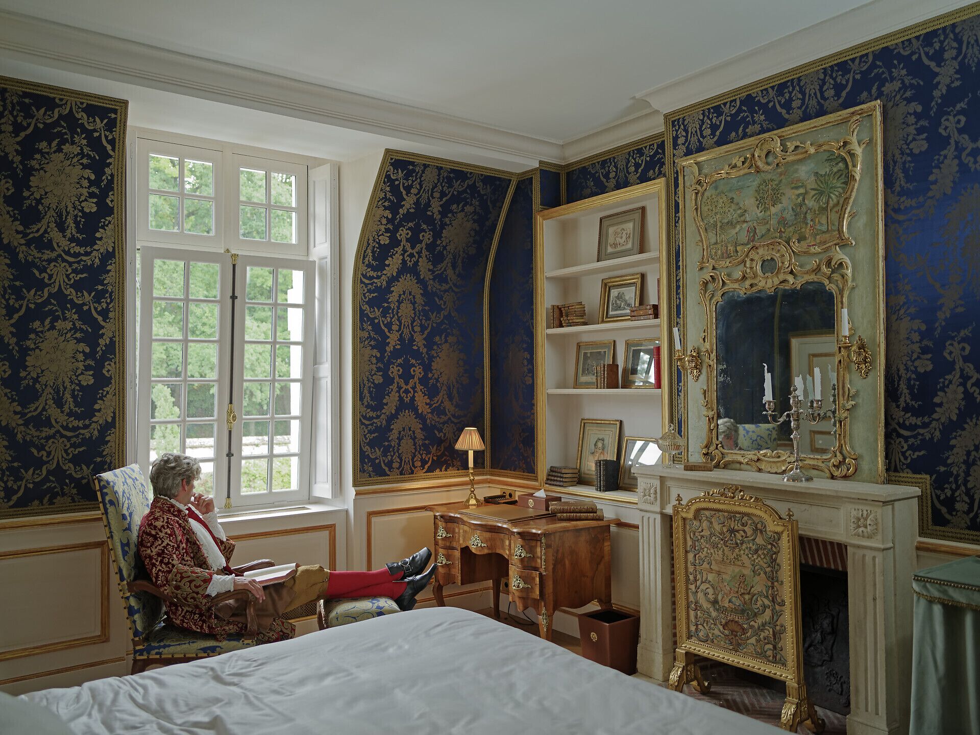 A room with a large bed, a desk, a chair, a fireplace, and a large mirror.