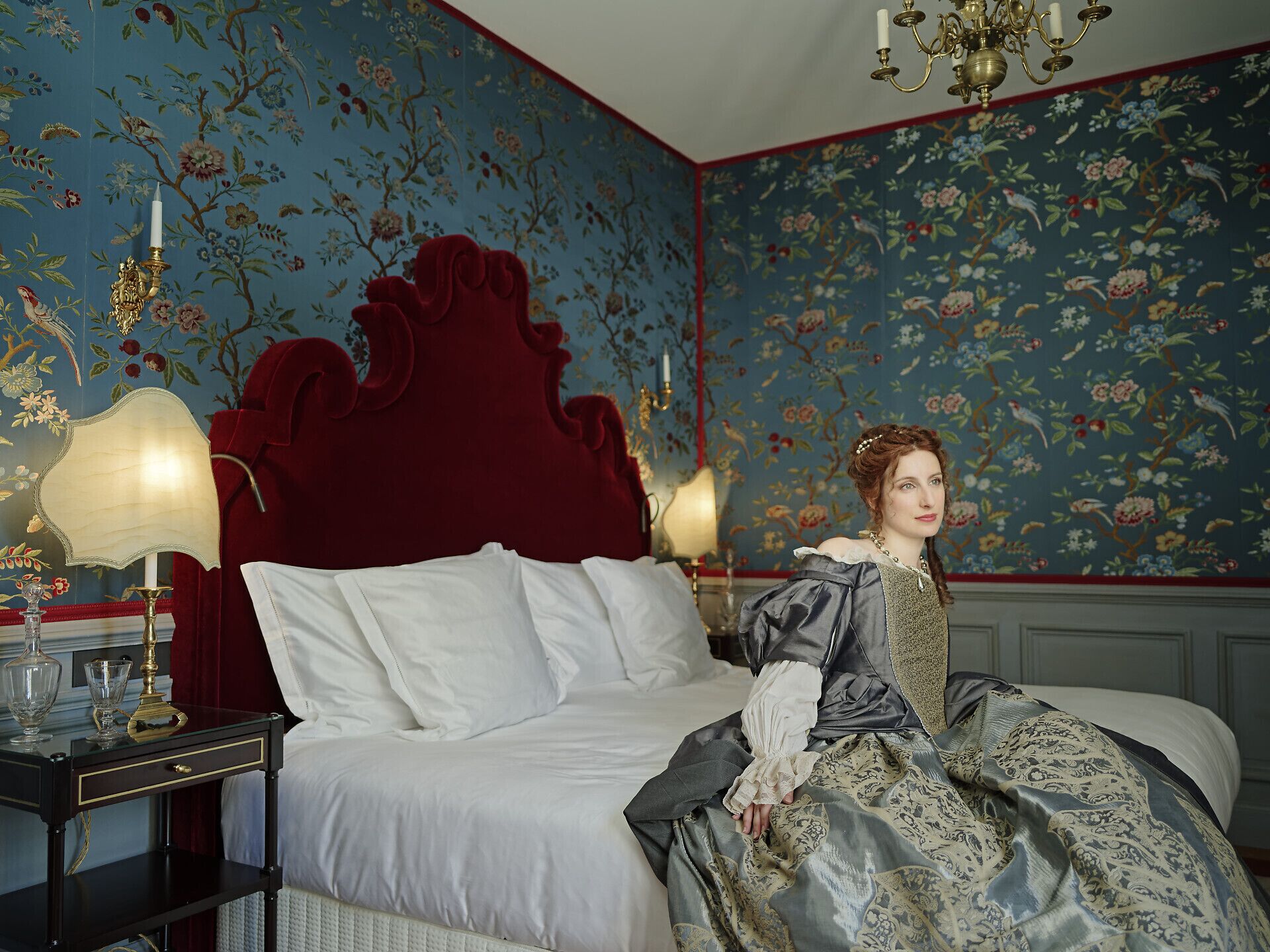 A bedroom with a large bed, ornate headboard, and floral wallpaper. There are two bedside tables with lamps and a mirror.