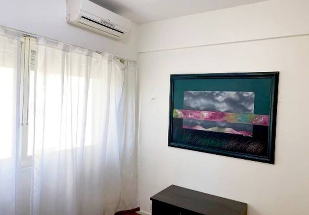 Ein Zimmer mit einem Fenster, das mit durchsichtigen Vorhängen bedeckt ist, einer Klimaanlage darüber und einem gerahmten abstrakten Gemälde an der Wand.