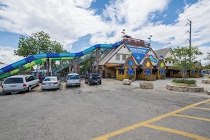 A water park entrance with a large blue and green slide, a wooden building with a sign reading 'WORLD'S BEST WATER PARK,' and several cars parked in front.