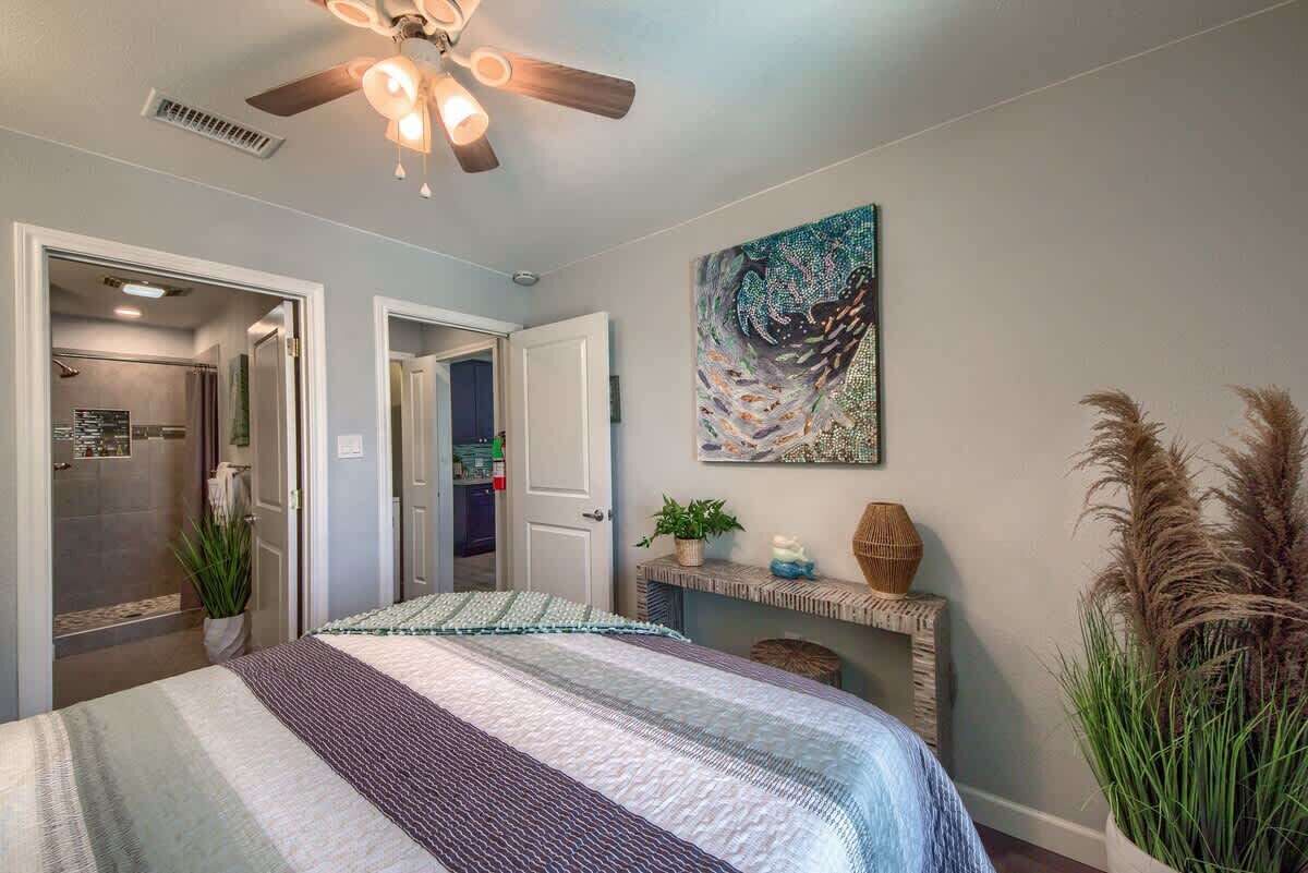 A bedroom with a bed, a ceiling fan, a painting, and a plant.