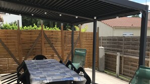 Une terrasse extérieure avec une table et des chaises couvertes, une clôture en bois et un sol carrelé.