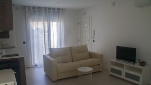 Ein Wohnzimmer mit einem beigen Sofa, einem kleinen runden Tisch, einem Fernseher auf einem weißen Schrank und einer Tür mit einem Telefon.