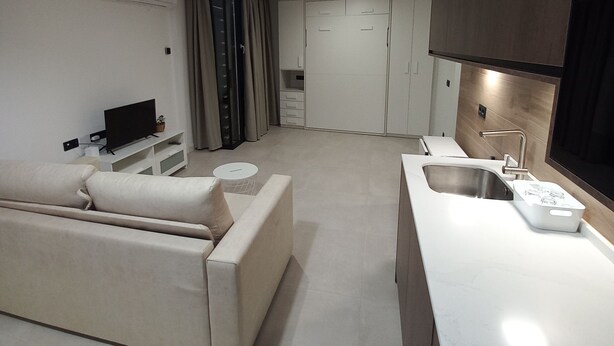 Eine moderne Küche mit Backofen, ein Essbereich mit weißem Tisch und Stühlen sowie ein Wohnzimmer mit Sofa und Fernseher.