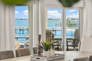 A dining area with a table set for two, featuring a tray with a potted plant and candle holders, overlooking a body of water and dock.