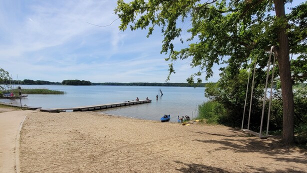 Une plage de sable avec une jetée qui s’avance sur un lac paisible, le tout entouré d’arbres et de verdure.