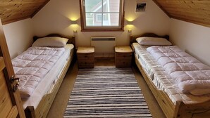 Une petite chambre mansardée, chaleureuse et accueillante, avec deux lits simples, un plafond en bois incliné, une fenêtre donnant sur une maison et un radiateur central.