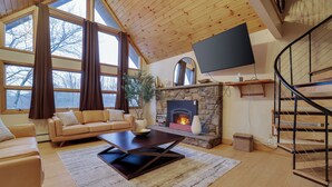 A cozy living room with a stone fireplace, a large window with curtains, a sofa, and a flat-screen TV mounted on the wall. - Paradise Pocono - Ultimate Arcade, Salt Pool and Hot Tub - Sleeps 14 (Cresco)