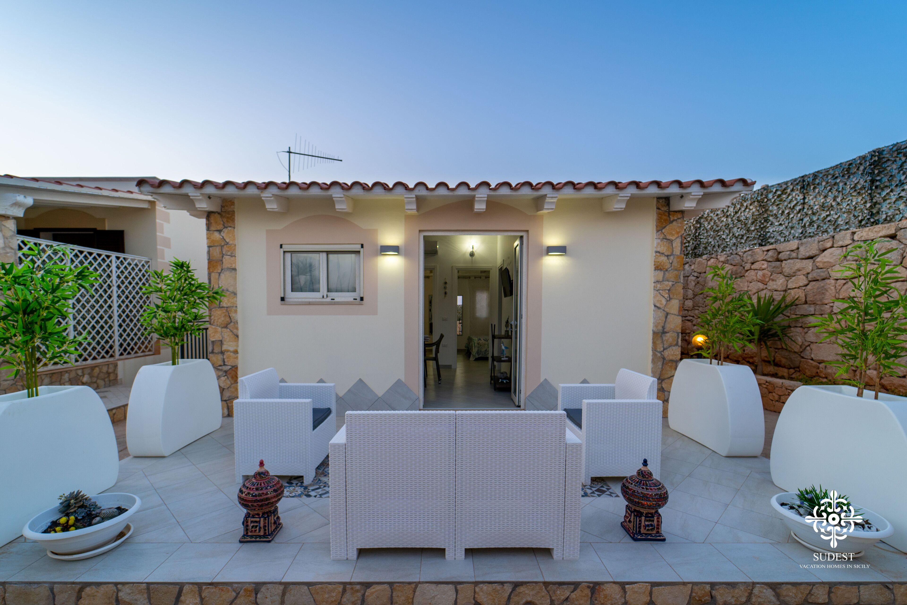 Un patio al aire libre con mobiliario de mimbre blanco, paredes de piedra y plantas en macetas.