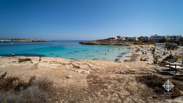 Vista costera con una playa arenosa, aguas azules cristalinas y edificios al fondo.