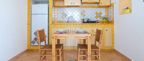 Una cocina compacta con refrigerador, armarios de madera, protector contra salpicaduras de azulejos y una mesa de comedor con sillas.