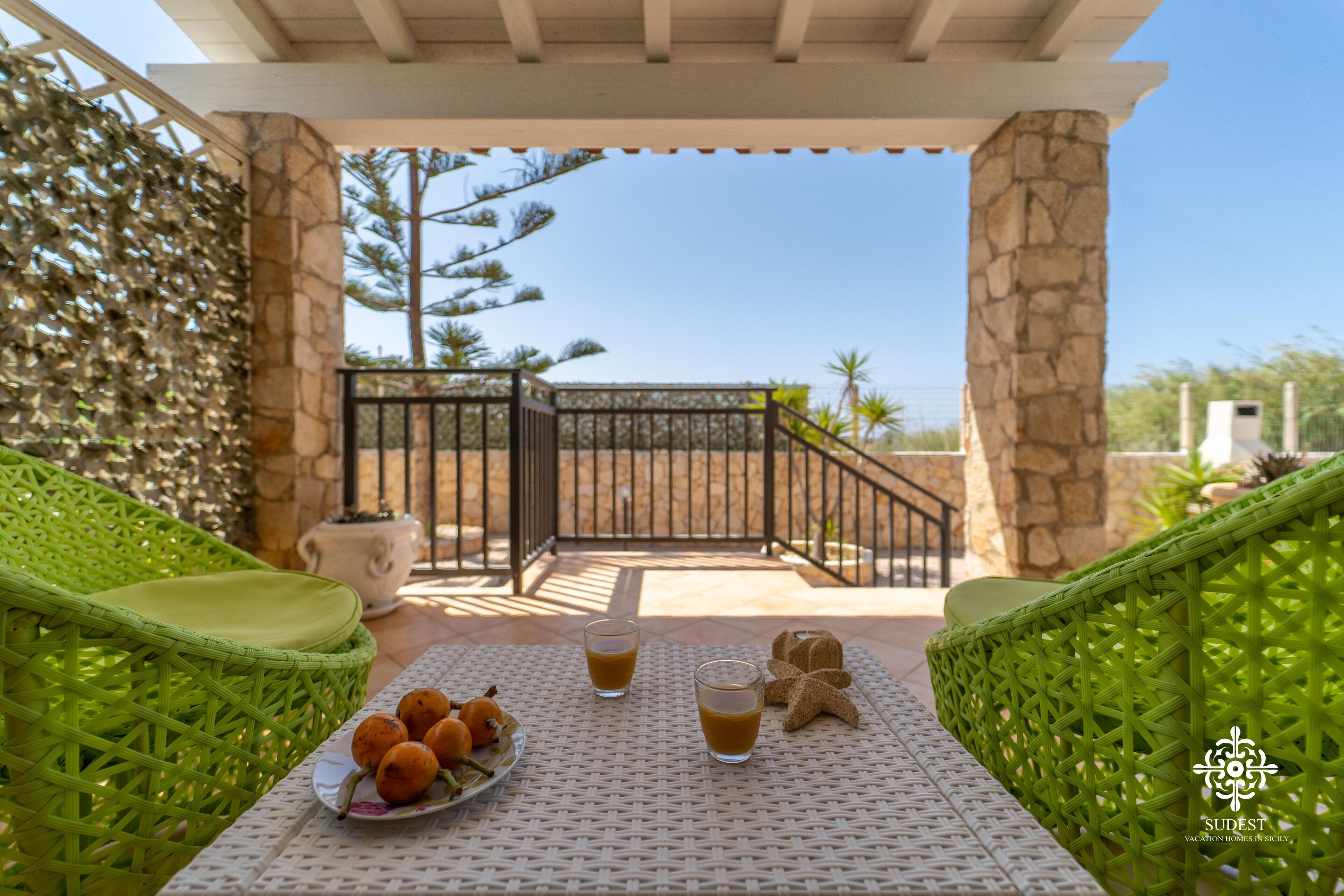 Un tavolo con due bicchieri di bevanda, un piatto con cibo e un biscotto a forma di stella, su un patio con sedie in rattan verde e un muro in pietra.