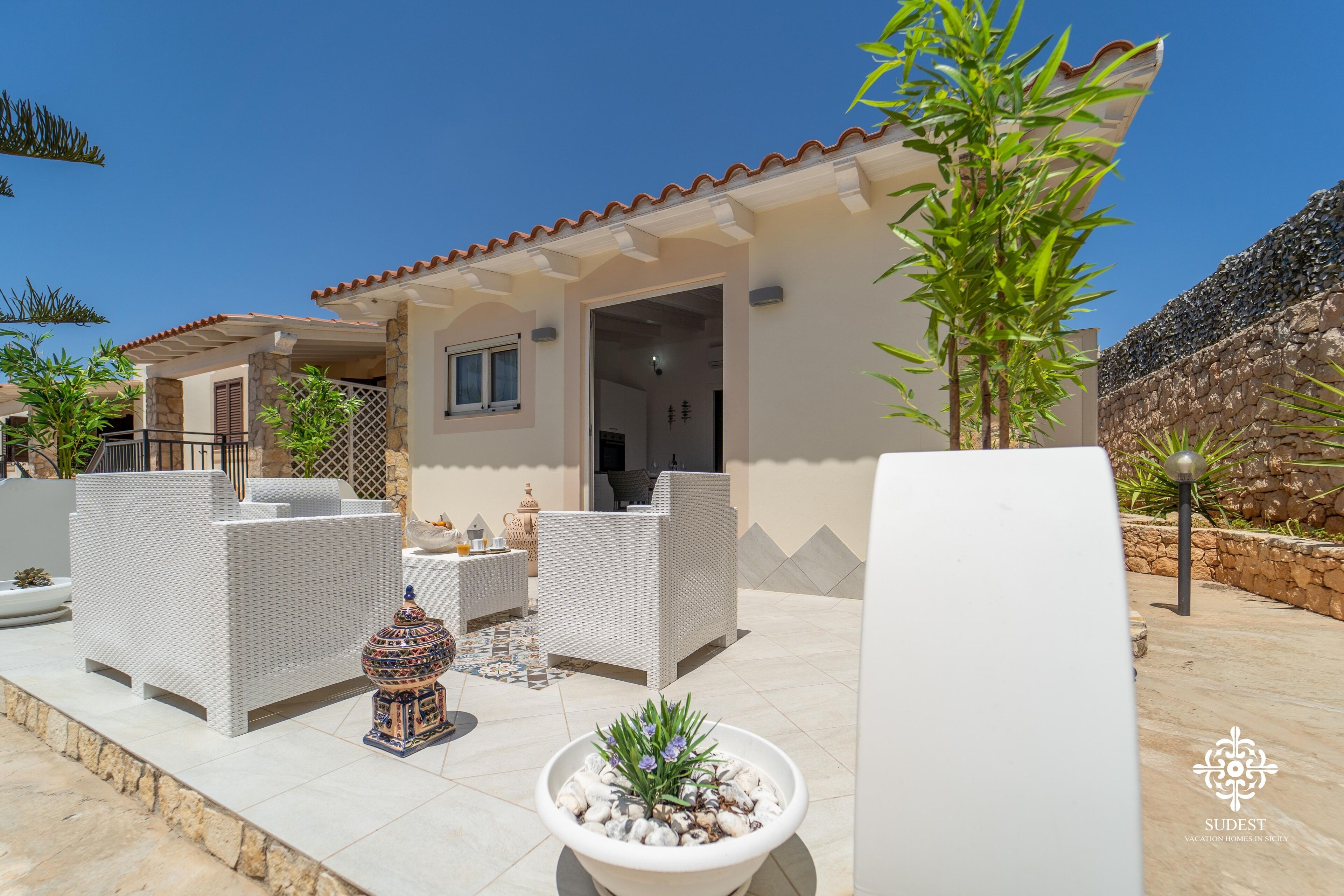 A modern villa with a patio area featuring wicker furniture, a decorative fountain, and a stone wall.