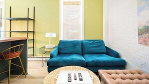 A living room with a teal sofa, a brown leather ottoman, a wicker coffee table with remote controls, a small side table with a lamp, and a metal shelf unit. - Tiny House Apt in Little Italy w OffStreet Parking (Wilmington)