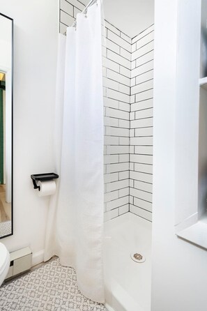 Une salle de bain avec une douche carrelée de carreaux blancs, des toilettes dotées d’un porte-serviettes en papier et un sol à motifs.