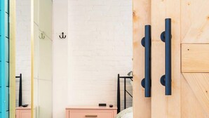 A bedroom with a white brick wall, a wooden wardrobe, a bed with a white duvet, a pink dresser, and a patterned rug. - Tiny House Apt in Little Italy w OffStreet Parking (Wilmington)