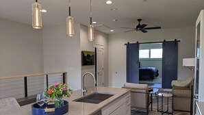 Une cuisine moderne avec un îlot central, un coin repas avec une petite table et des chaises, et une chambre visible à travers une porte coulissante.