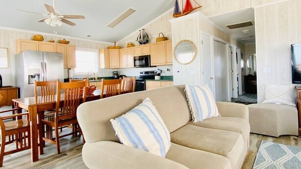 A living room with a beige sofa, wooden dining table, and chairs, a kitchen in the background, and a ceiling fan. - *Beach House!!*Views!!*Steps to Ocean!*Amenities! (Oak Island)