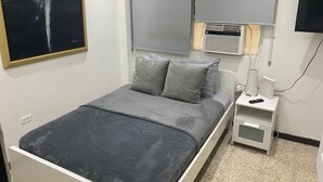 A bedroom with a bed, pillows, a nightstand, a lamp, a TV, and an air conditioning unit.