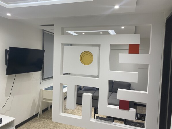 A modern living room with a flat-screen TV mounted on the wall, a white entertainment unit with a circular decorative piece, and a grey sofa with a red accent piece.