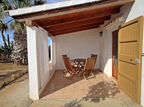 Um alpendre coberto com uma mesa e cadeiras de madeira, uma porta e uma parede branca.