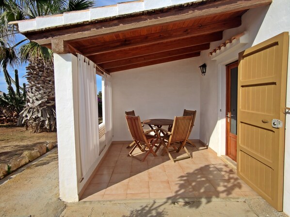 Une terrasse couverte avec une table et des chaises en bois, une porte et un mur blanc.