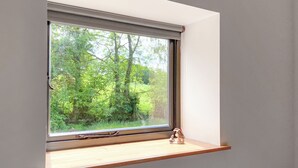 Une fenêtre donnant sur une forêt, une petite étagère et une vue partielle sur l’intérieur d’une pièce.