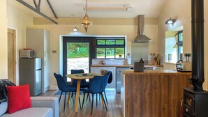 Une cuisine moderne avec un îlot central, un coin repas avec des chaises bleues, une cuisinière intégrée et une vue sur la verdure à l’extérieur.