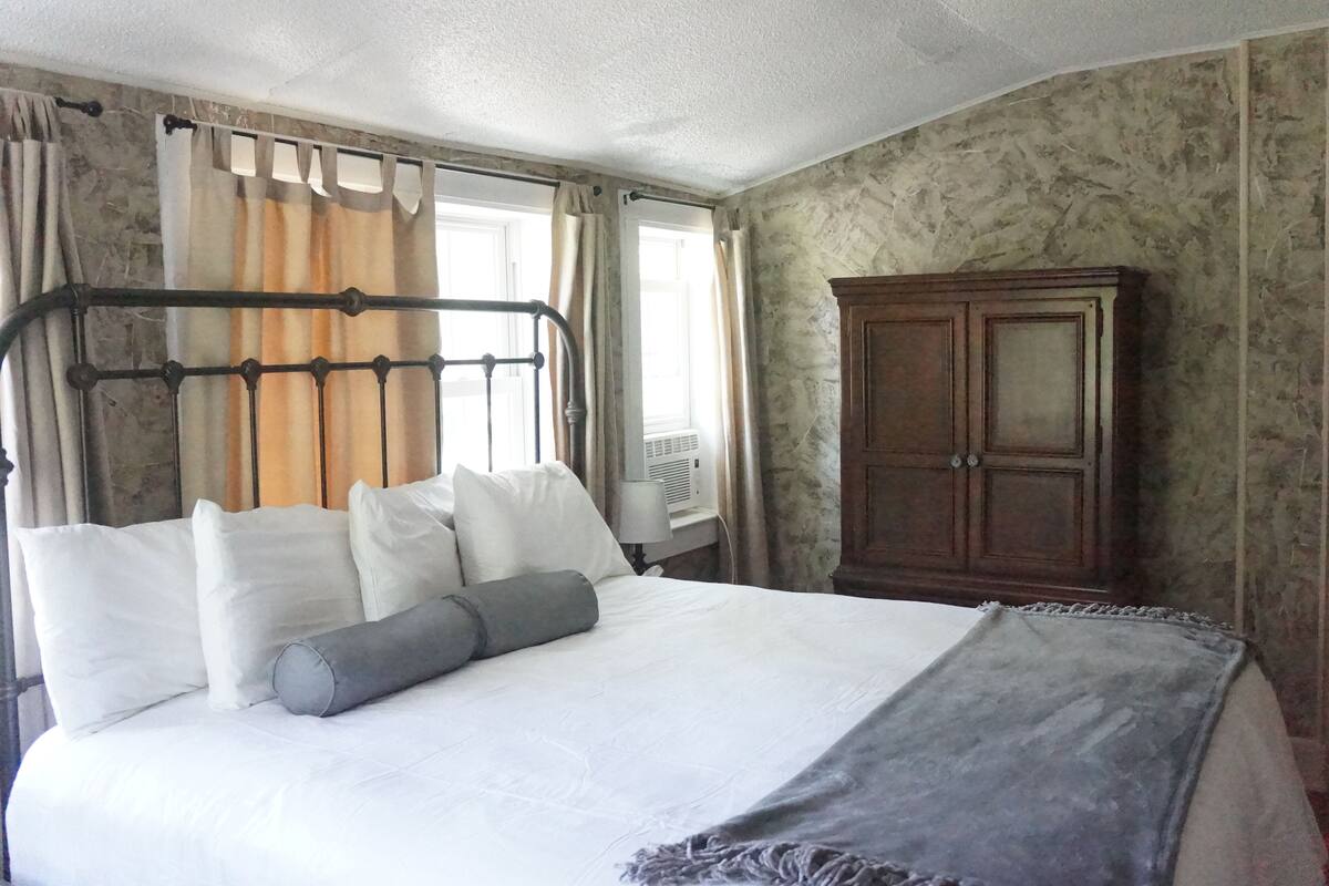 Ein Schlafzimmer mit einem Bett, Kissen, einer Decke, einem Holzschrank und Vorhängen.