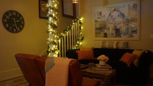 A living room with a brown armchair, a black sofa, a coffee table with books, a clock, and a painting of a barn on the wall. - The Frogtown Inn (Canadensis)